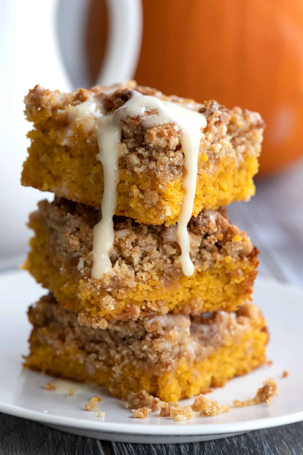 Squares of Keto Pumpkin Crumb Cake stacked on a white plate with glaze dripping down the side.