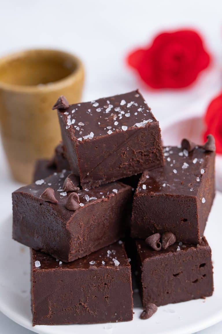 Creamy keto fudge piled up on a white plate with red flours in the background.