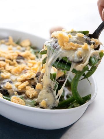 A spoon lifting out some Keto Green Bean Casserole from the dish, with gooey cheese on top.