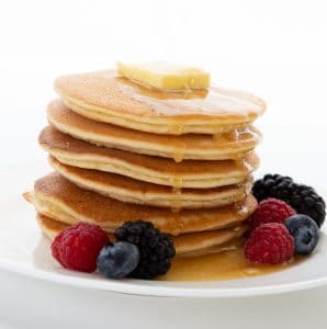 A stack of keto pancakes on a white plate, with butter on top and syrup dripping down. Fresh berries are scattered around.