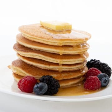 A stack of keto pancakes on a white plate, with butter on top and syrup dripping down. Fresh berries are scattered around.