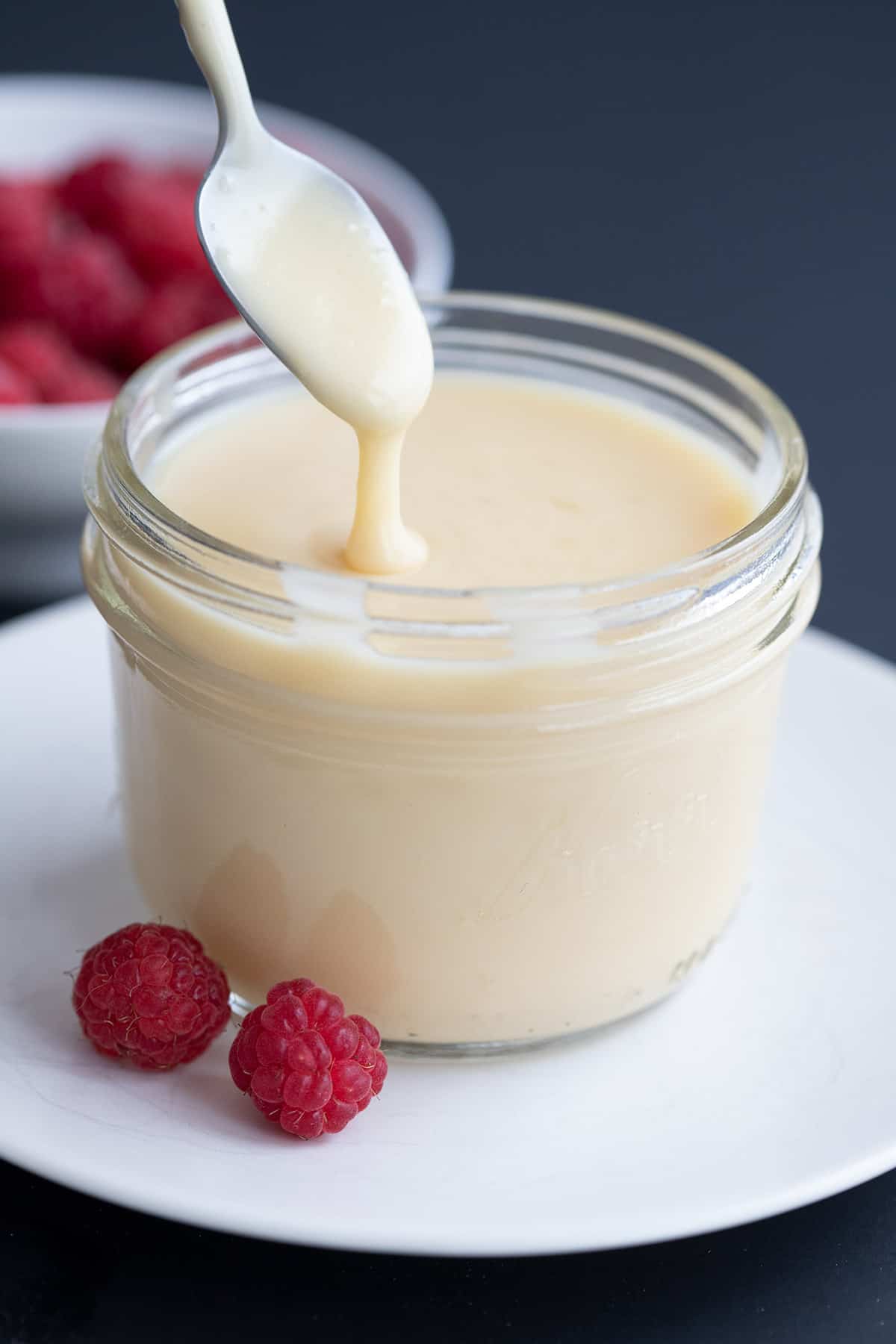 A jar of Keto Condensed Milk on a white plate with raspberries around it.