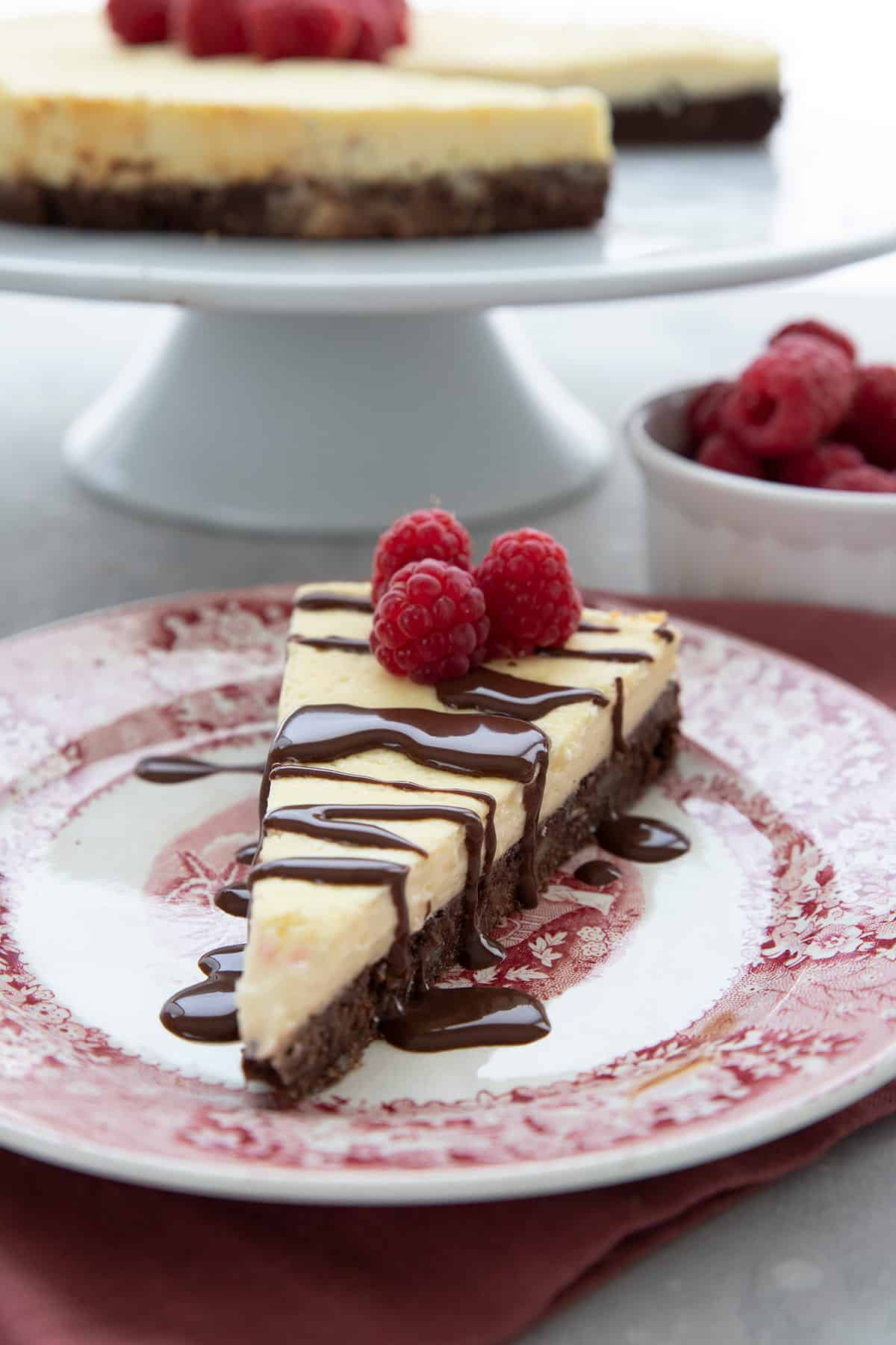 A slice of Keto Brownie Cheesecake sits on a plate in front of a cake stand.