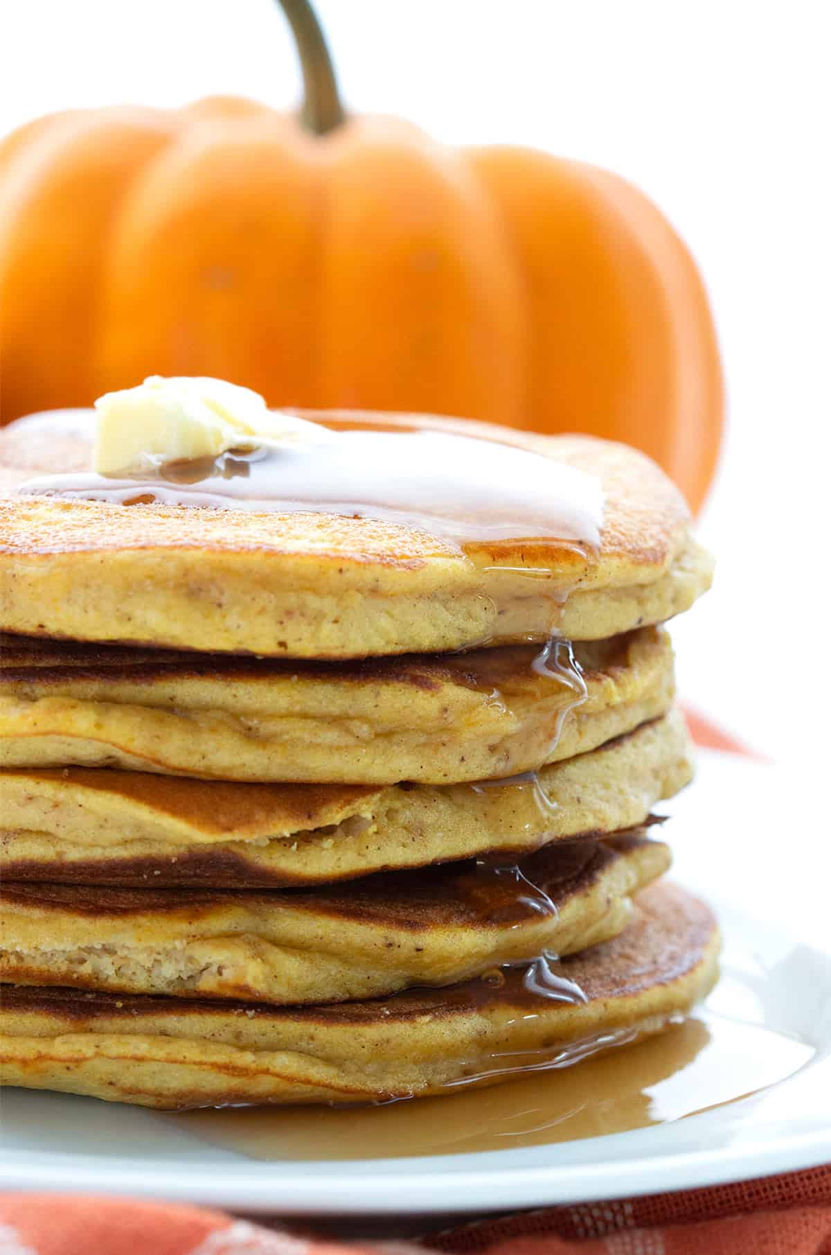 Close up shot of a stack of Keto Pumpkin Pancakes with syrup dripping down, in front of a small pumpkin.