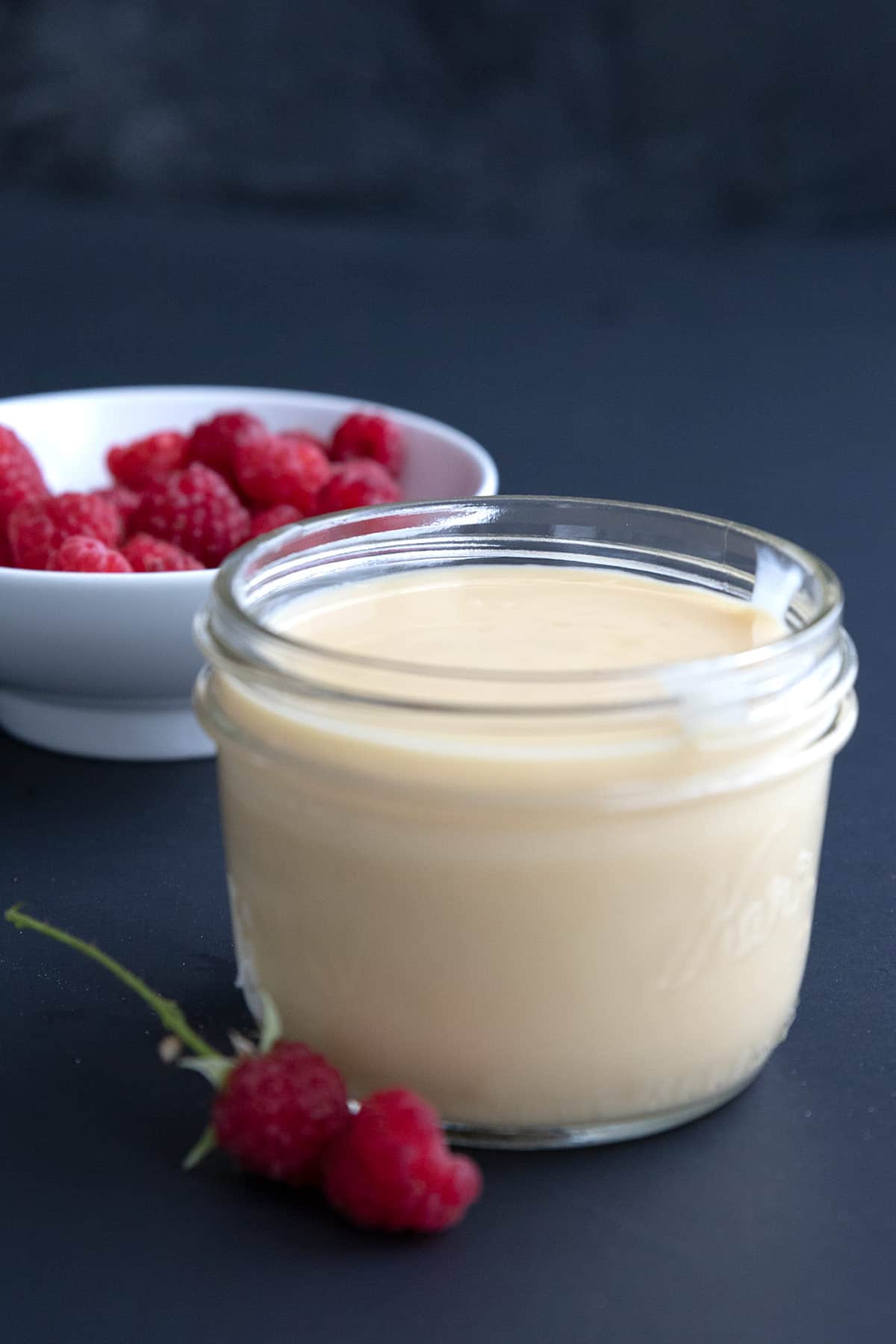 A jar of keto sweetened condensed milk on a dark blue background with a bowl of raspberries in behind.