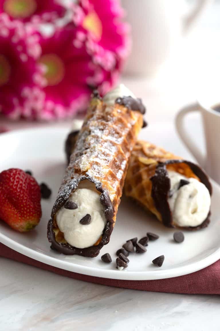 Two keto cannoli on a white plate with a strawberry, and pink flowers in the background.