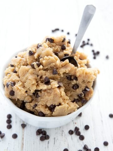 Edible keto cookie dough piled in a white bowl with a small spoon, and chocolate chips strewn around.