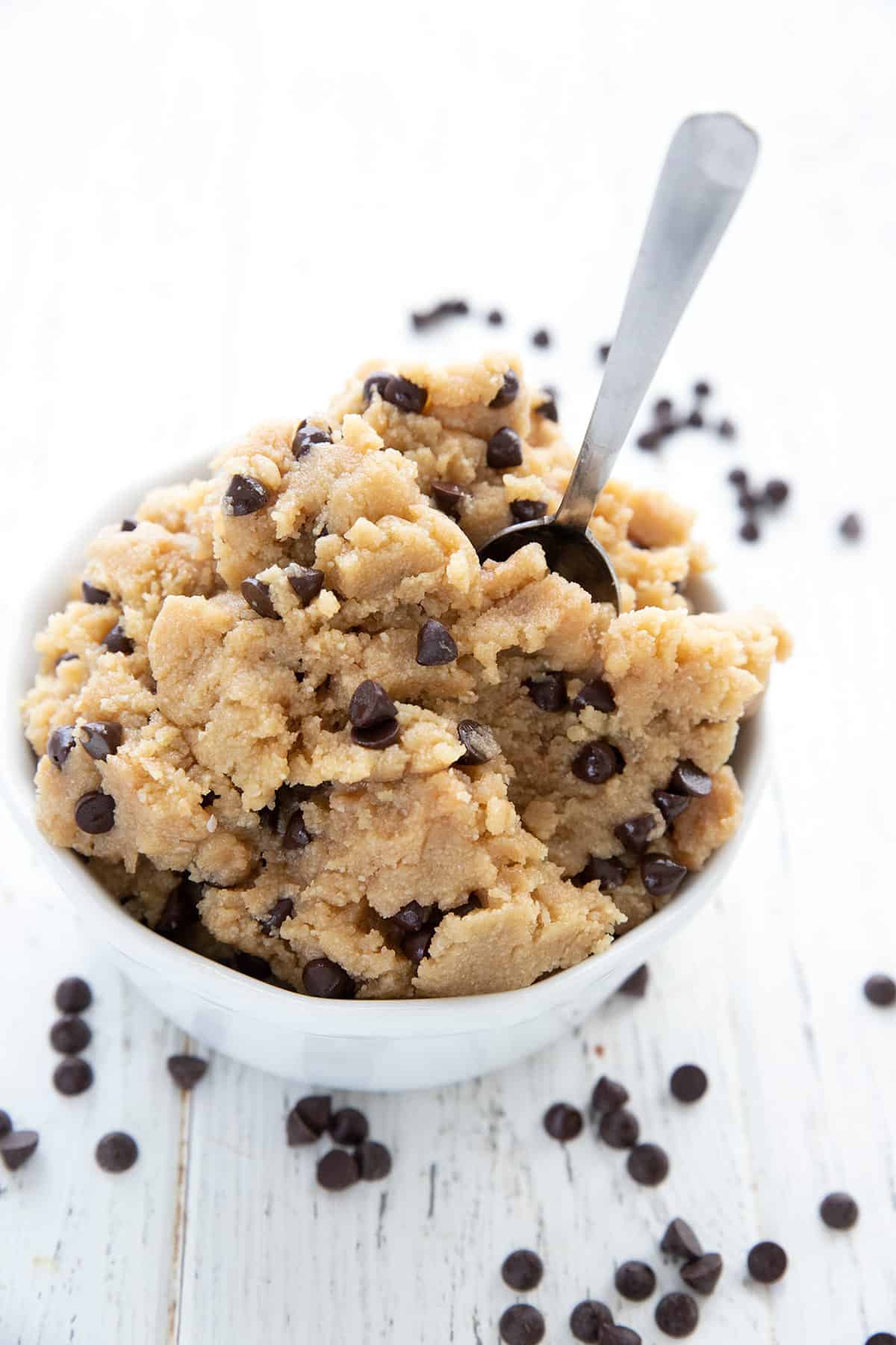 Edible keto cookie dough piled in a white bowl with a small spoon, and chocolate chips strewn around.