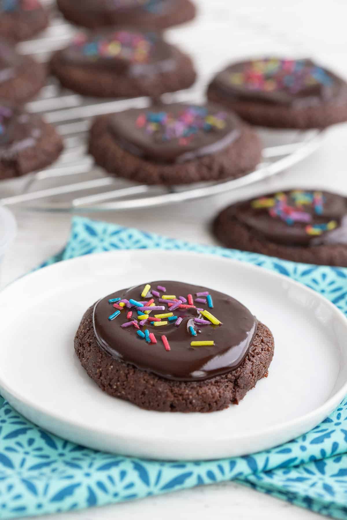 A keto chocolate cookie with glaze and sprinkles sits on a white plate in front of more cookies on a cooling rack.