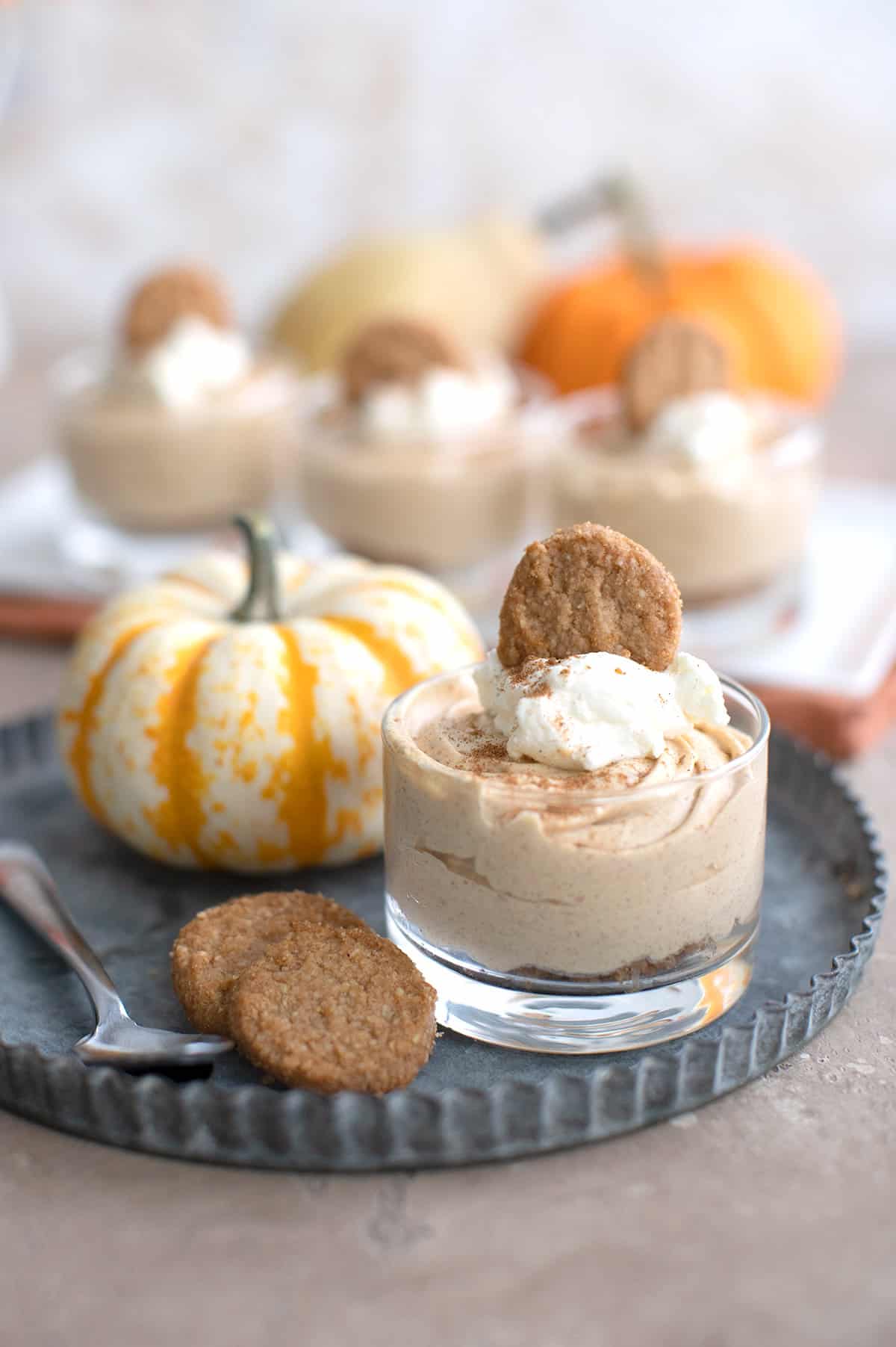 A jar filled with No Bake Keto Pumpkin Cheesecake on a pewter plate, with keto ginger cookies and a pumpkin.