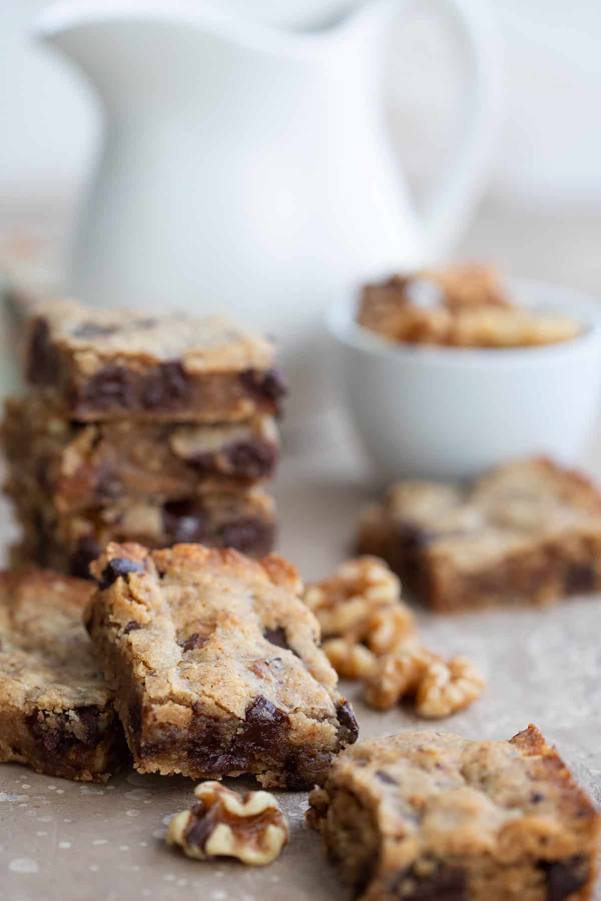 Walnut blondies and walnuts strewn around a table with a white pitcher in the background.