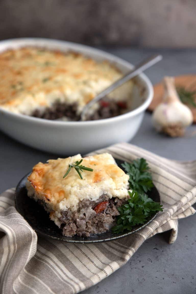 A slice of Keto Shepherd's Pie on a black plate over a striped gray napkin, with the rest of the pie in the background.