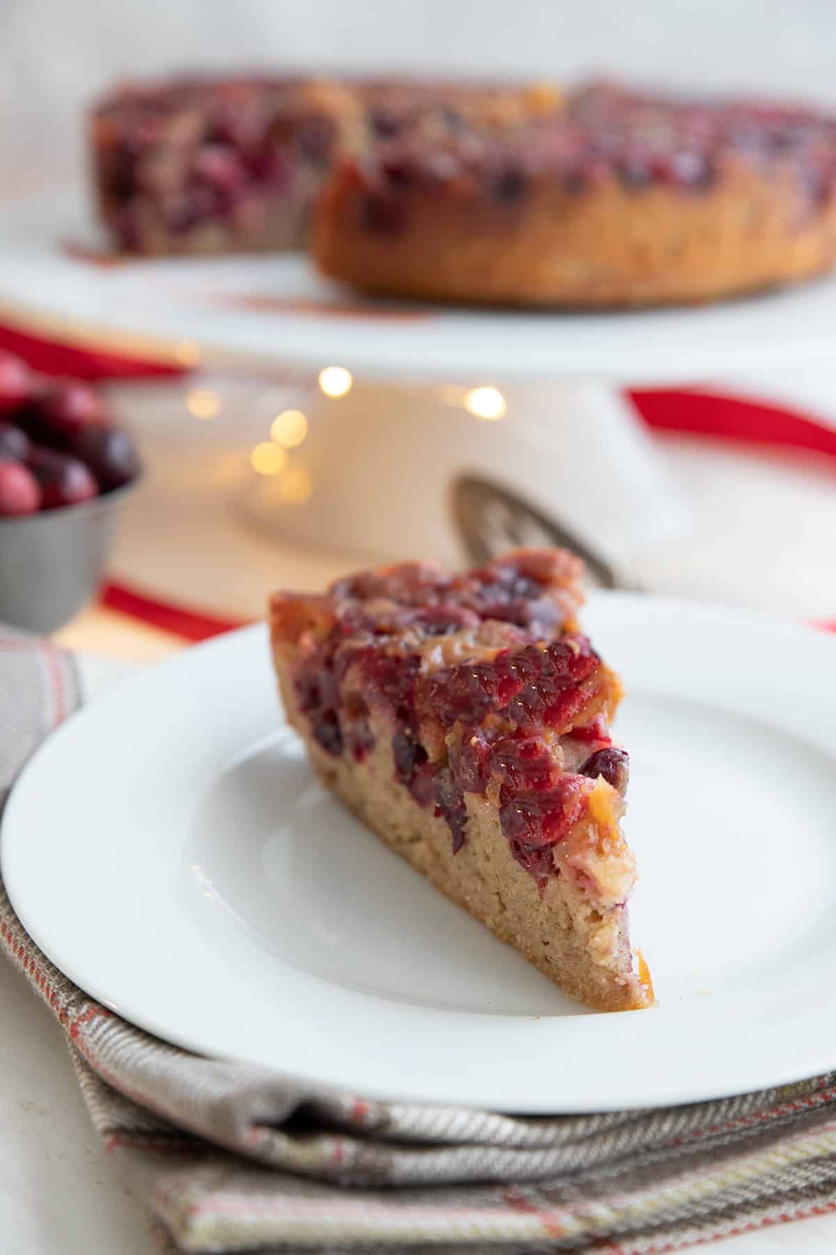 A slice of cranberry upside down cake on a white plate over a grey plaid napkin.