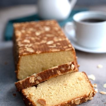 A loaf of almond flour cake on a gray concrete table with sliced almonds strewn around.