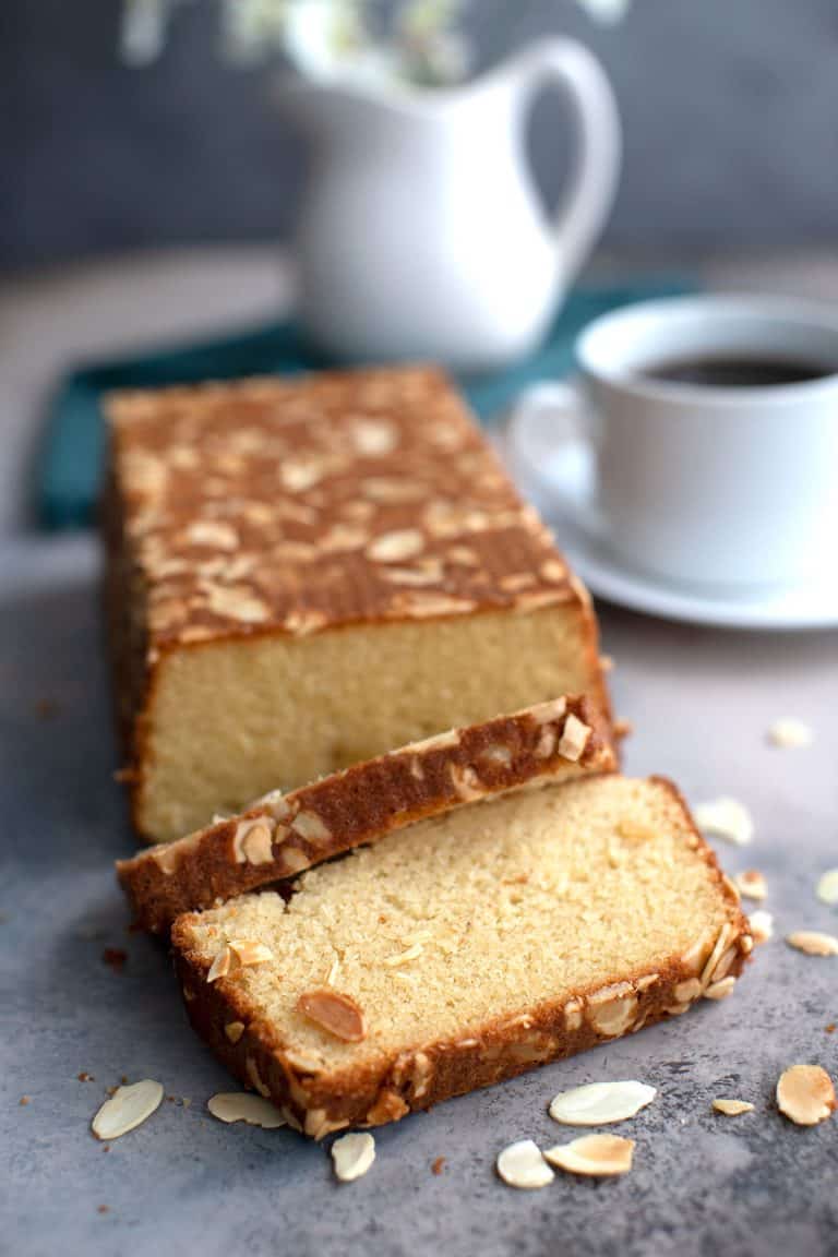 A loaf of almond flour cake on a gray concrete table with sliced almonds strewn around.