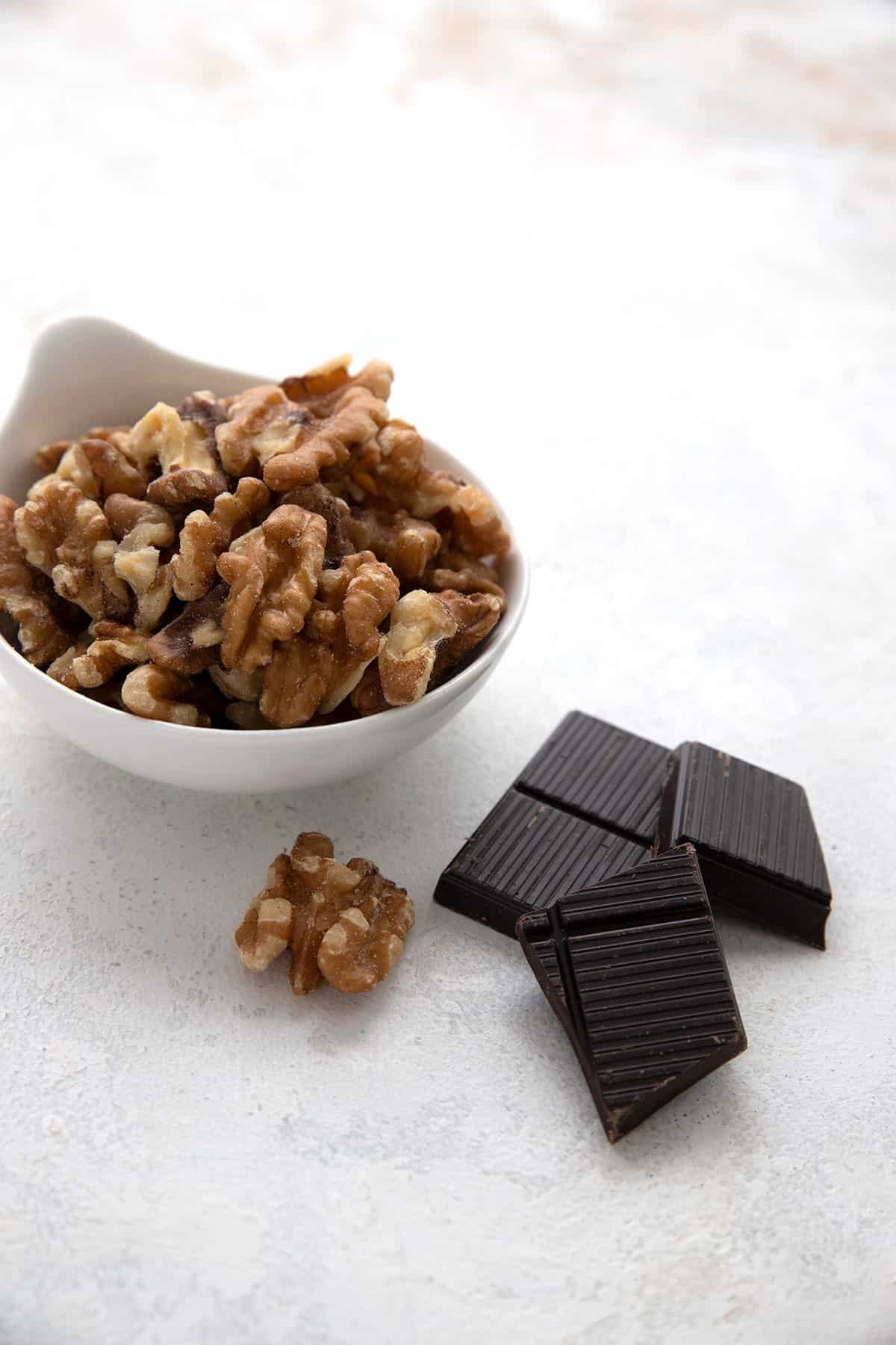 A bowl of walnuts and a few pieces of dark chocolate on a marble table.