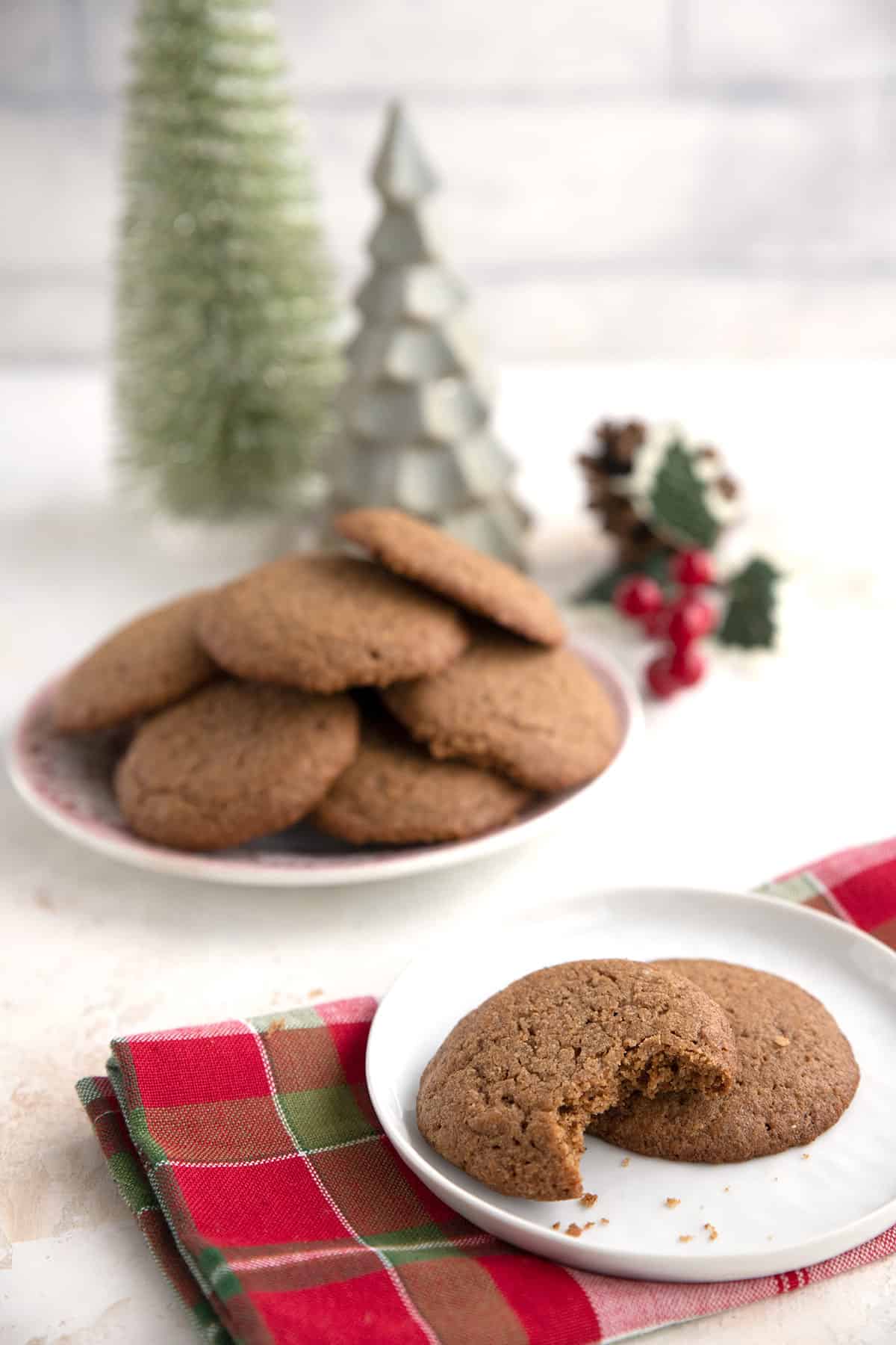 Two keto ginger molasses cookies on a white plate in front of more cookies and some holiday decor.