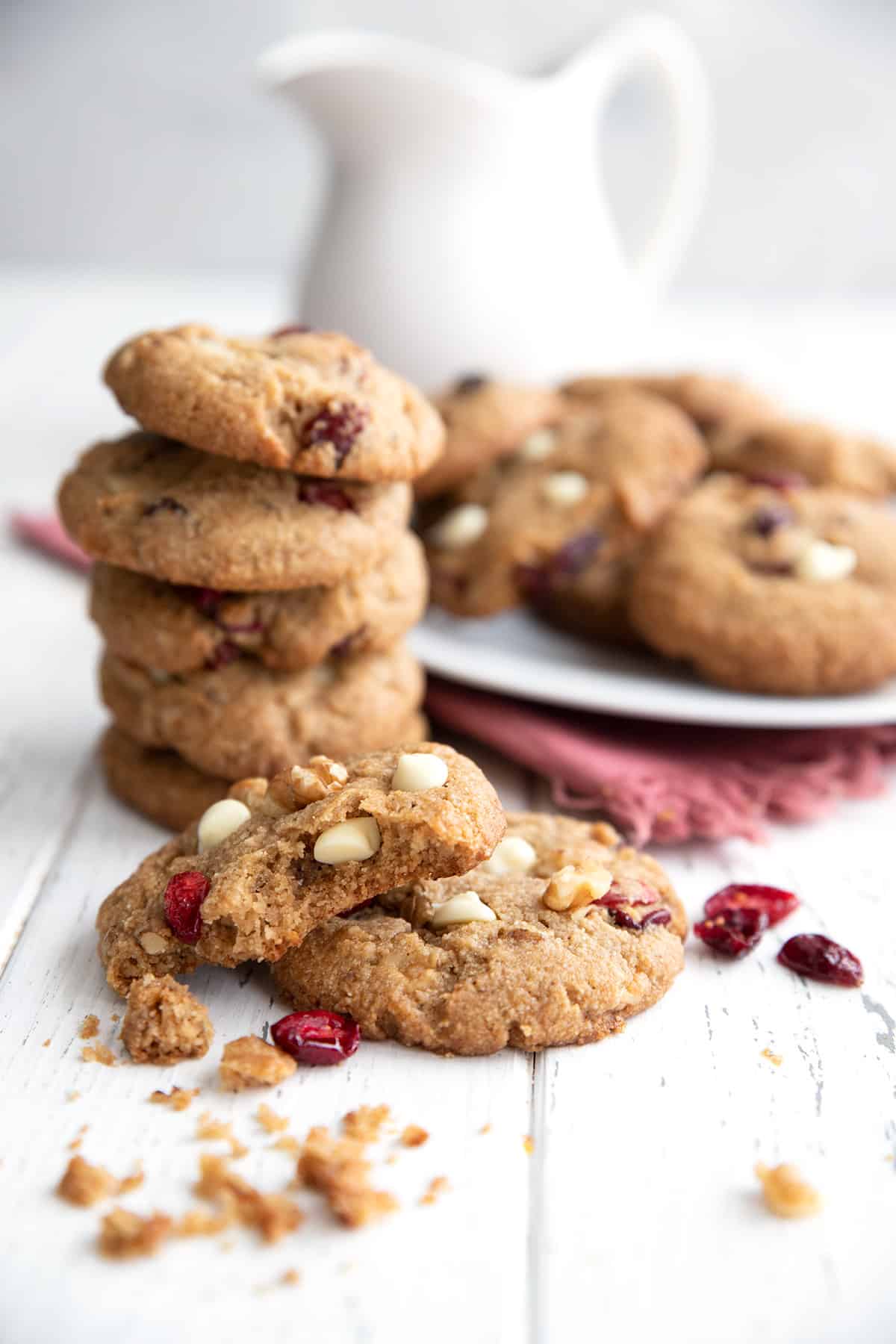 Two Keto White Chocolate Cranberry Cookies sit on a white table with a plate of more cookies in the background.