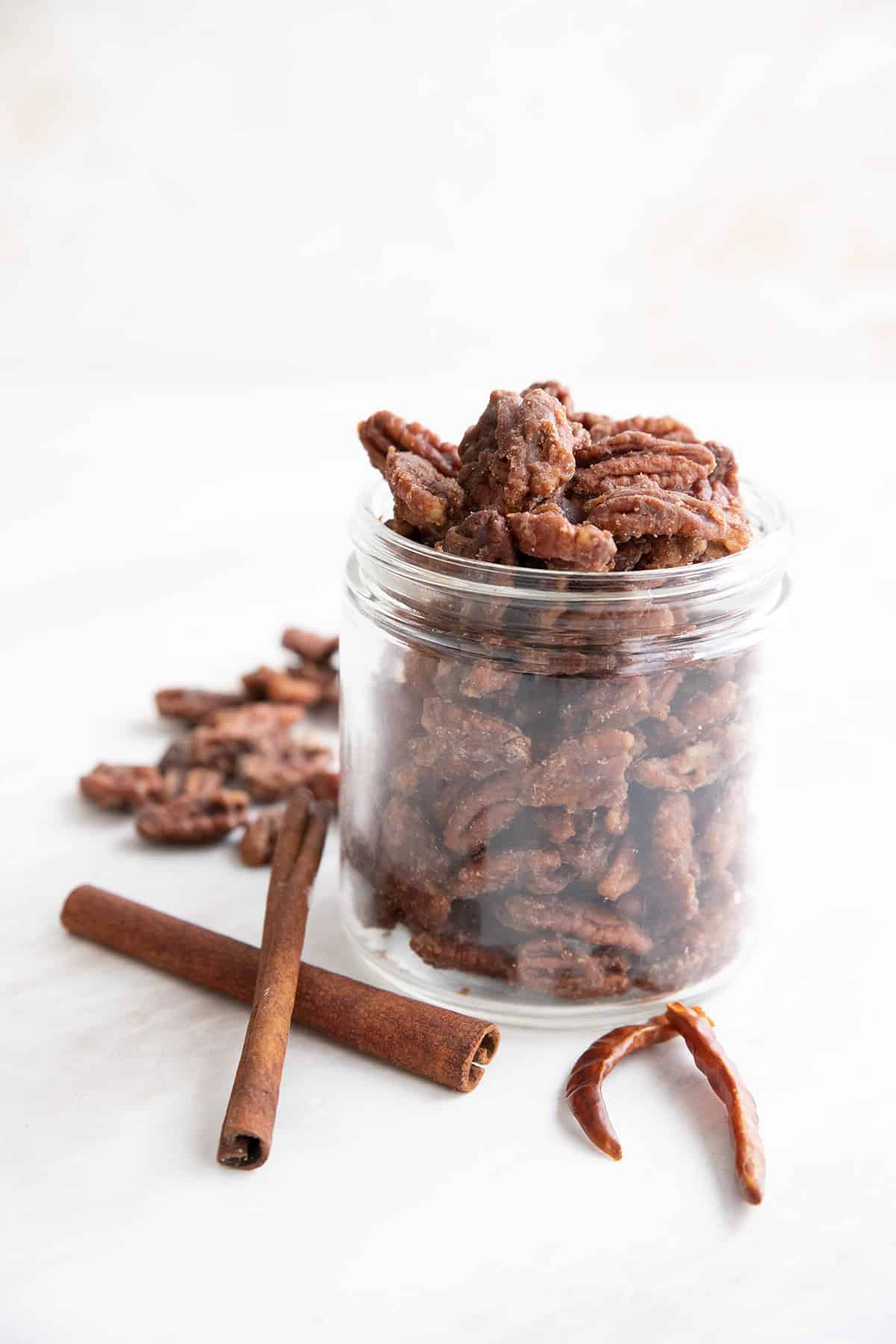 A jar of keto candied pecans with cinnamon sticks and dried chilis in front.
