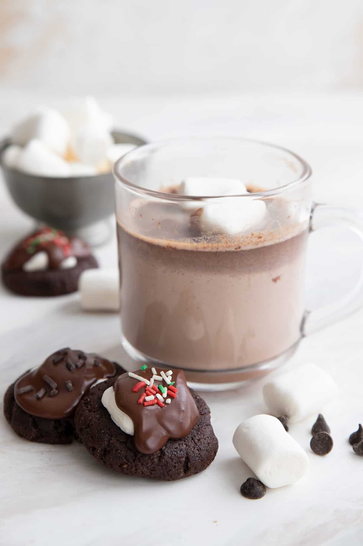 Two keto hot chocolate cookies beside a cup of sugar free hot chocolate with marshmallows.