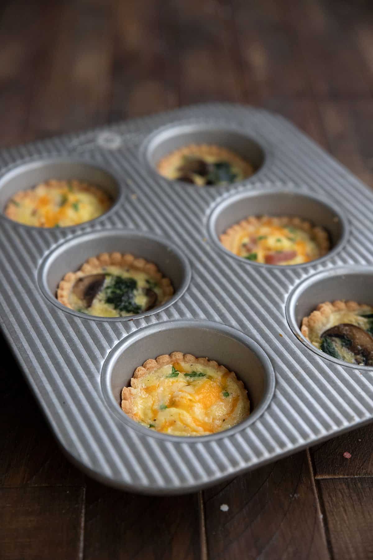 Two flavors of mini quiche in a metal muffin pan on a brown wooden table.
