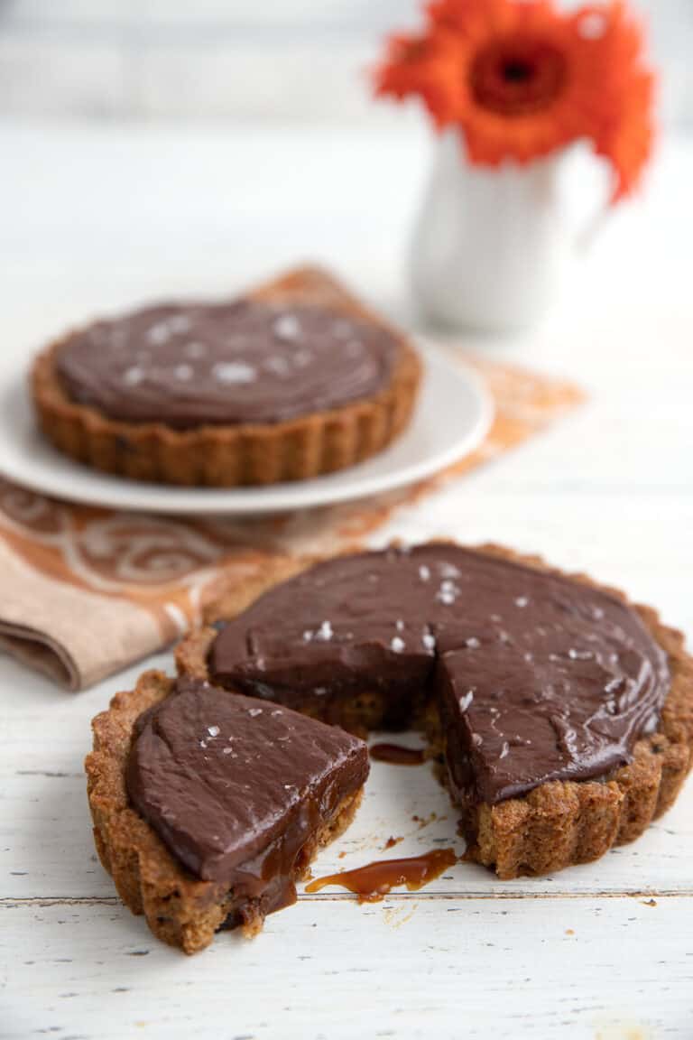 Two small keto salted caramel tarts on a white table, with a slice taken out of the front tart.