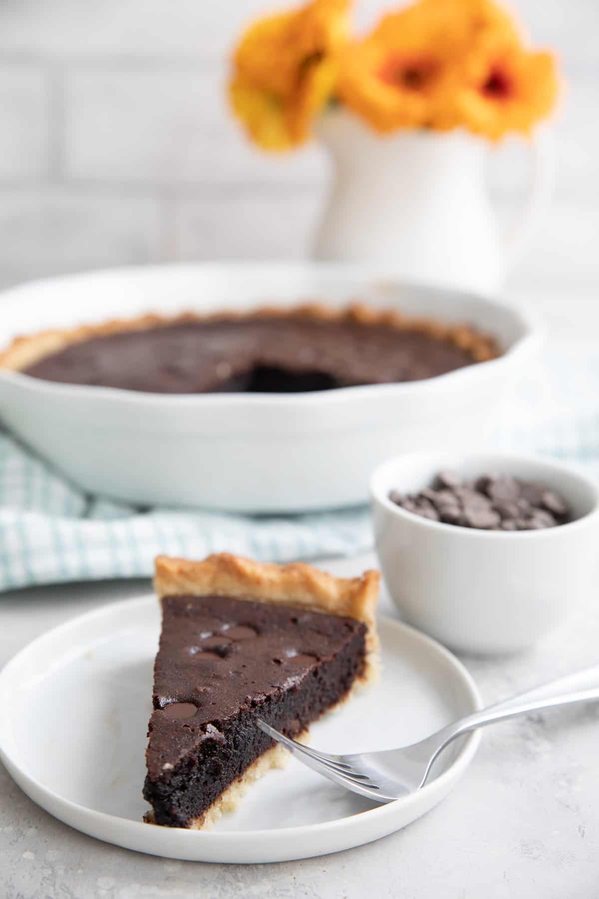 A slice of brownie pie on a white plate with the rest of the pie in the background.