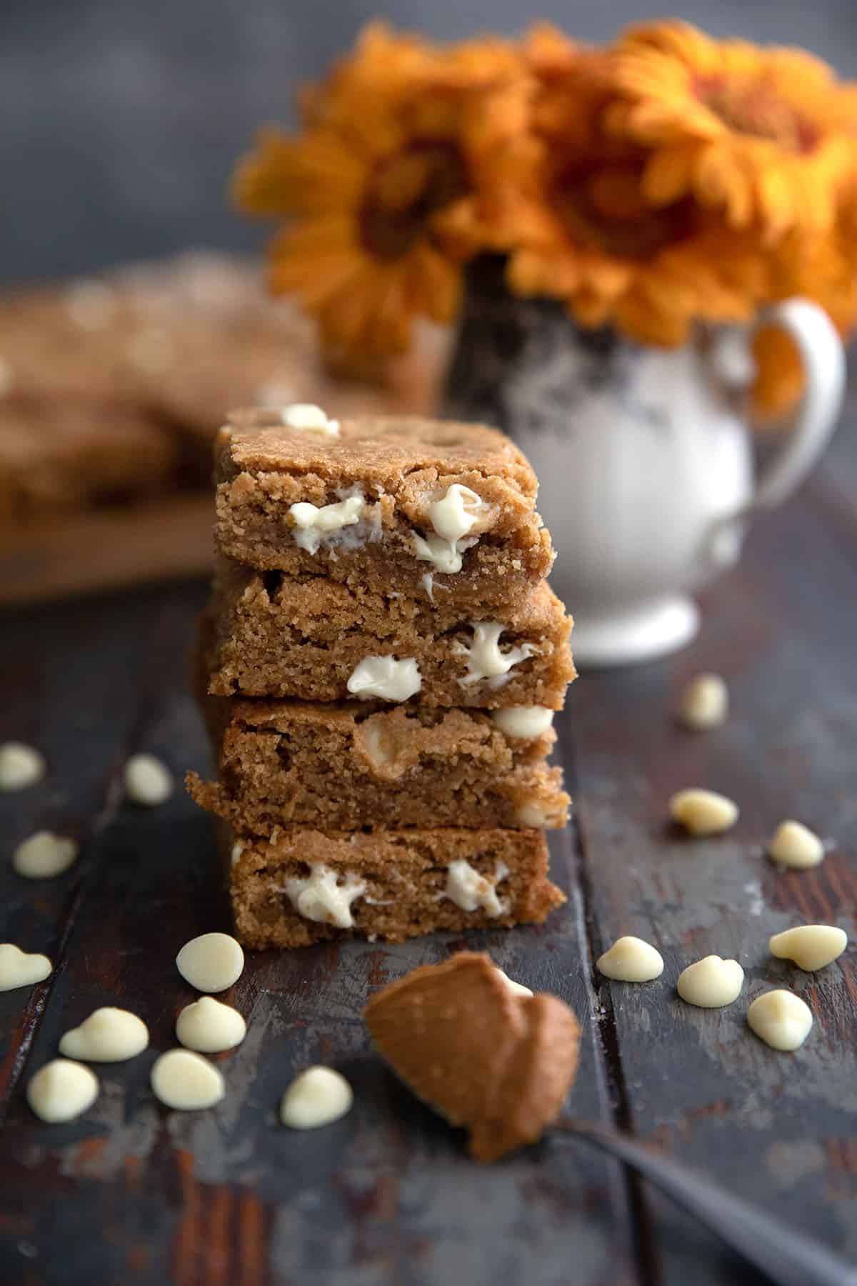 A stack of keto cookie bars with a scoopful of keto cookie butter and some white chocolate chips in front. A vase of orange gerbera daisies in the background.