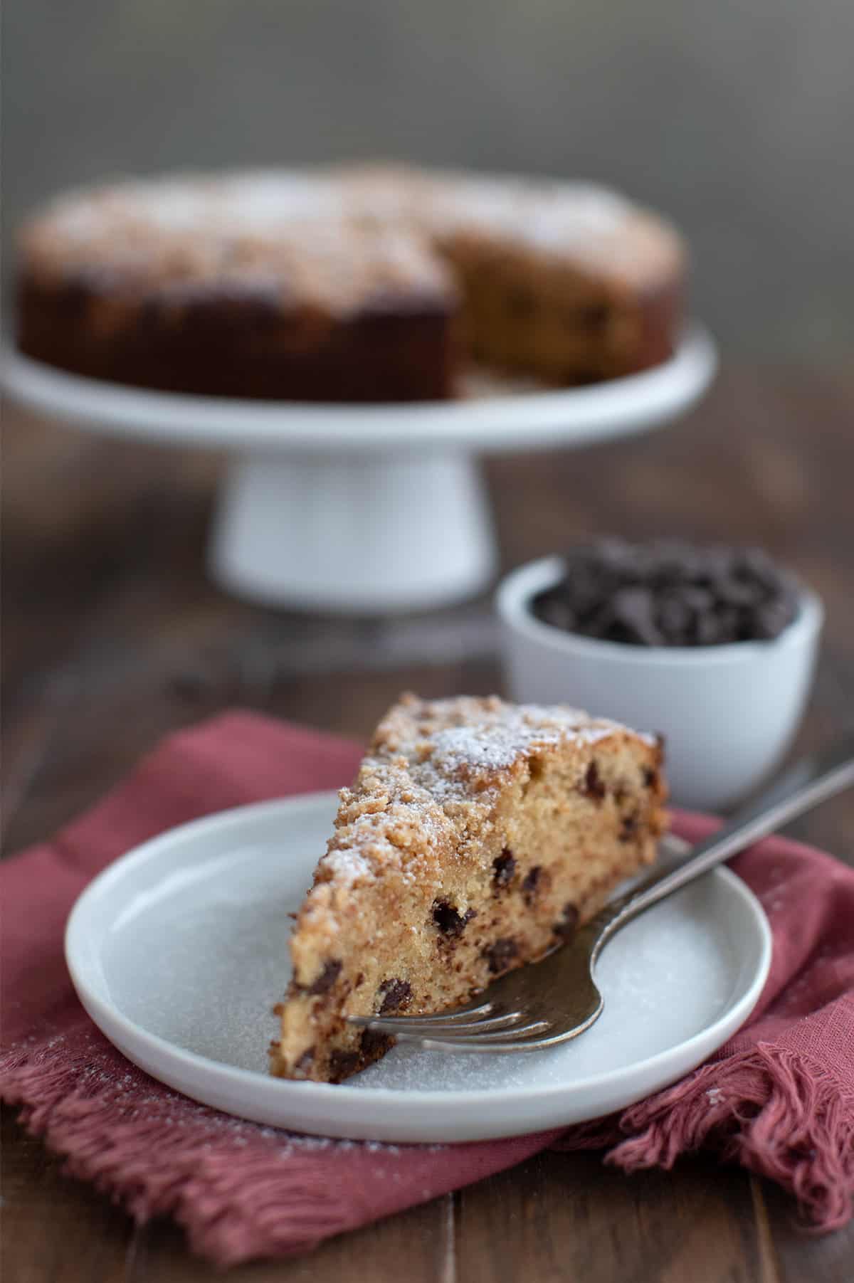 A slice of keto sour cream coffee cake on a white plate with a bowl of chocolate chips in the background.