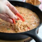 A hand dipping red pepper into cheesy sausage dip.