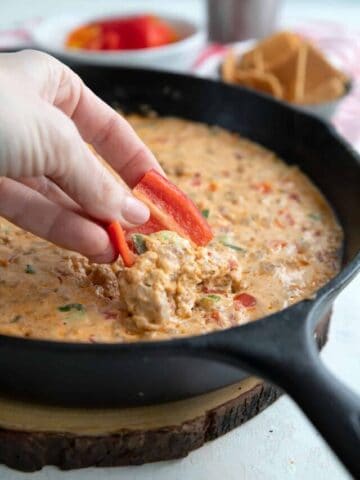A hand dipping red pepper into cheesy sausage dip.