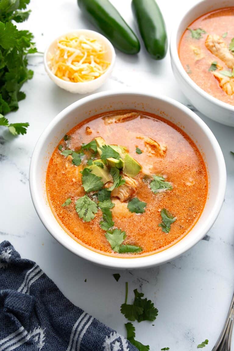 Two bowls of Chicken Enchilada soup on a white marble table top with cheese and jalapenos in the background.