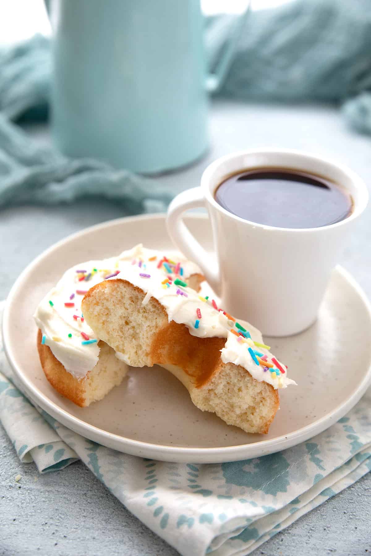 A vanilla protein donut broken open to show the inside on a white plate with a cup of coffee.