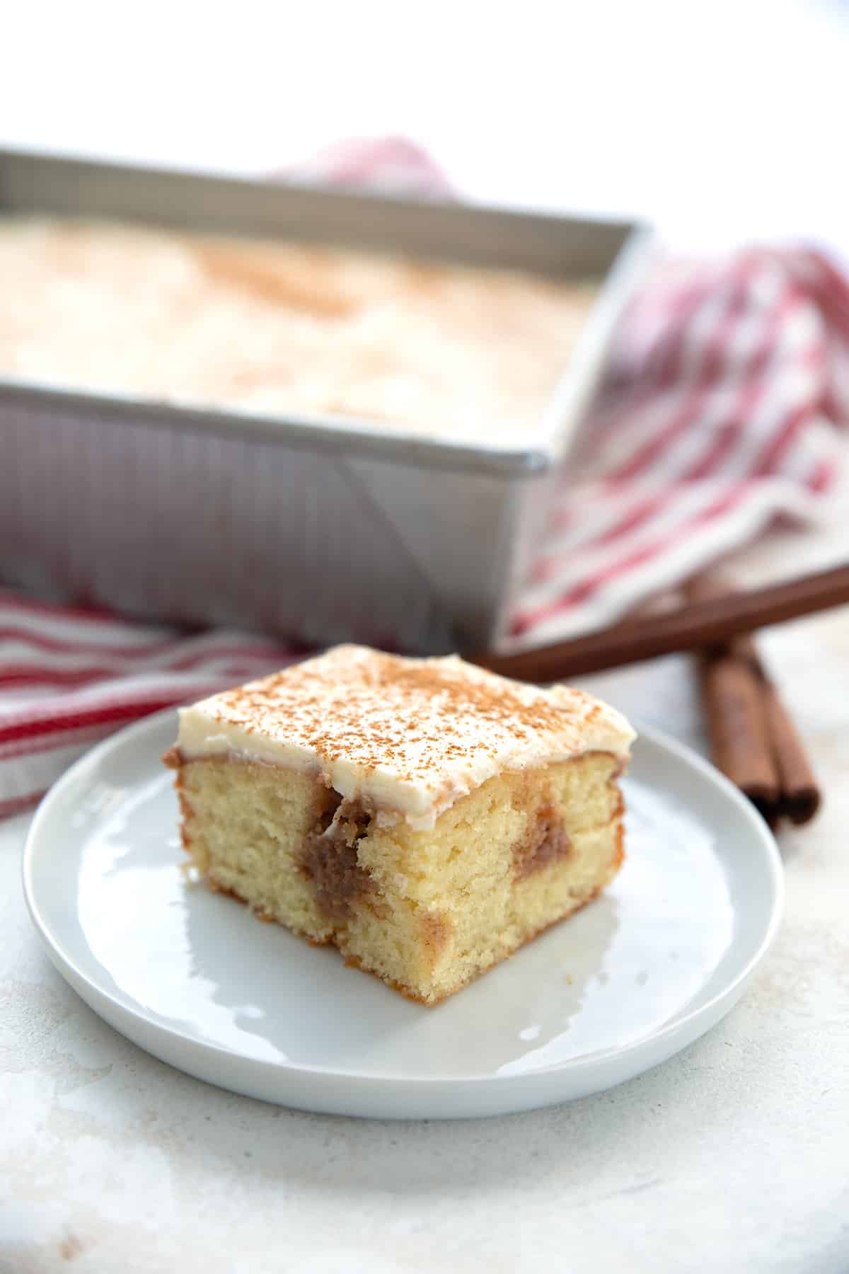 A slice of cinnamon roll poke cake on a white plate in front of the pan of cake.