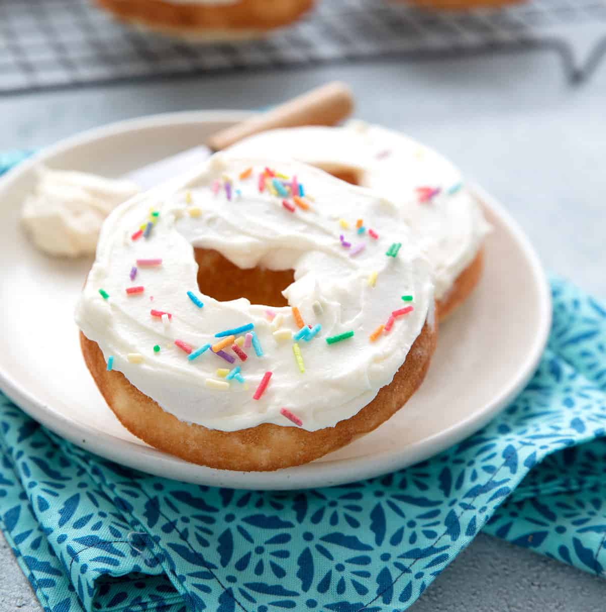 Two Vanilla Keto Protein Donuts sitting on a white plate over a teal patterned napkin.