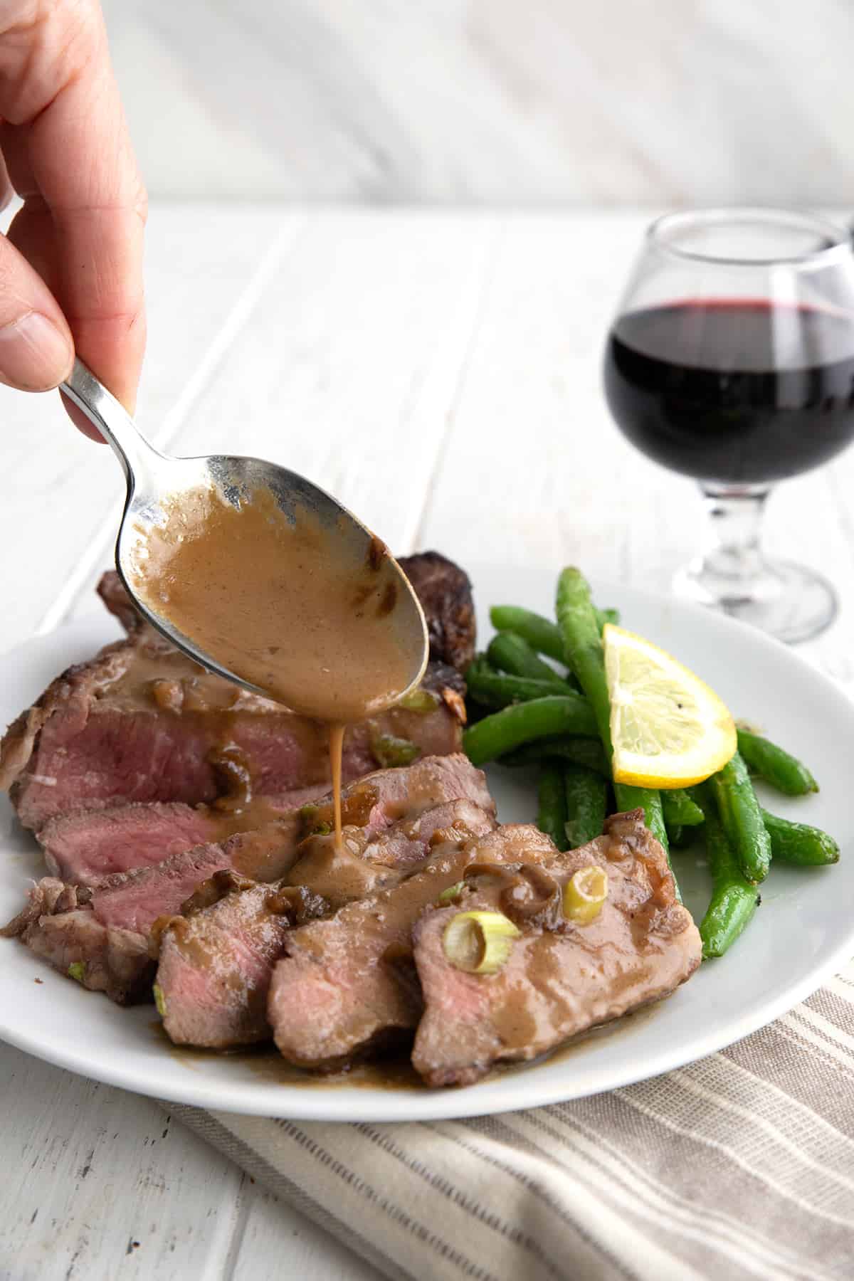 A spoon pouring sauce over steak Diane with a glass of wine in the background.