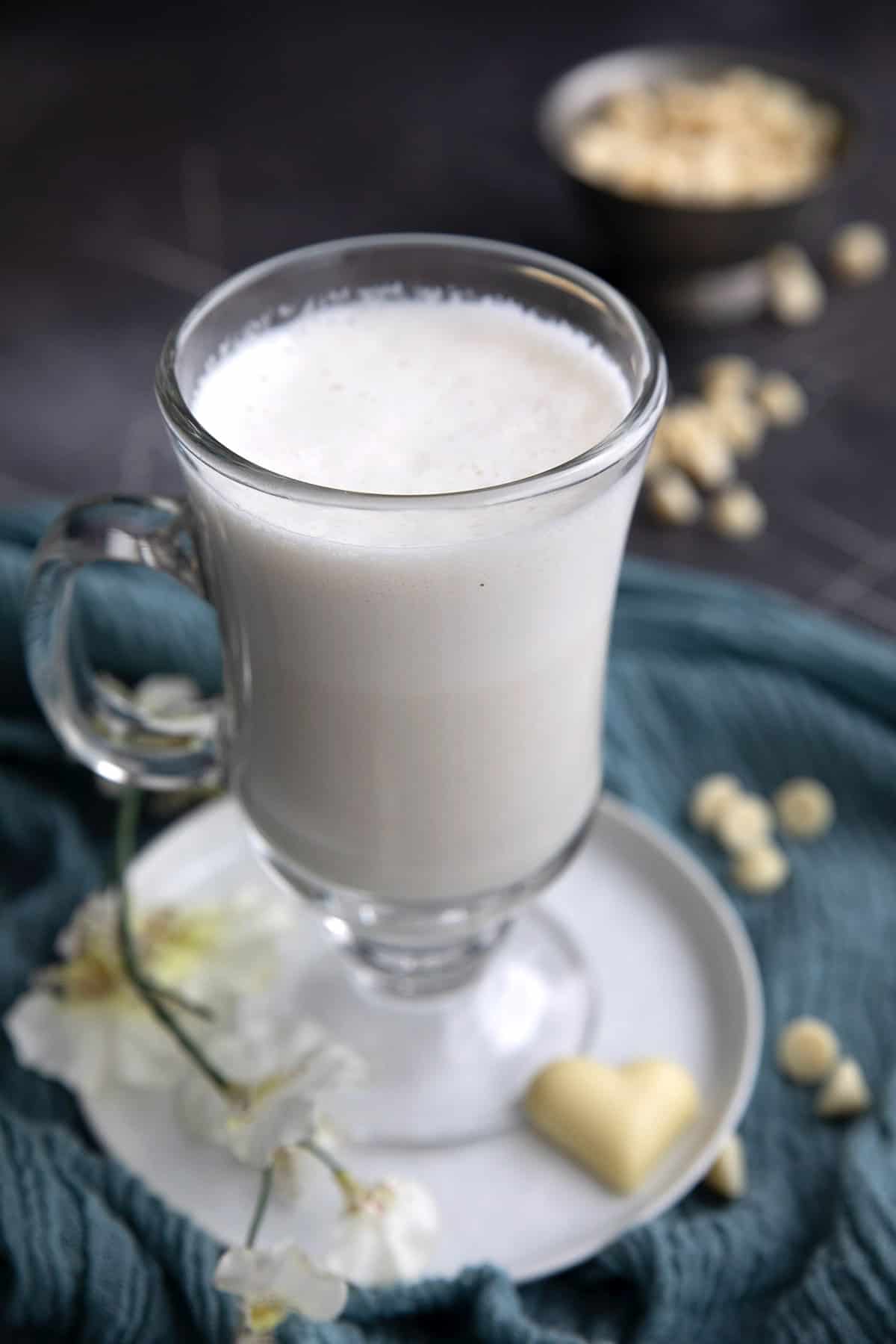 A glass mug filled with white hot chocolate with a bowl of white chocolate chips in the background.