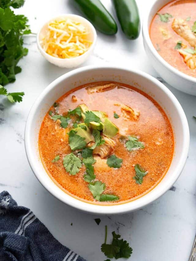 Two bowls of Chicken Enchilada soup on a white marble table top with cheese and jalapenos in the background.