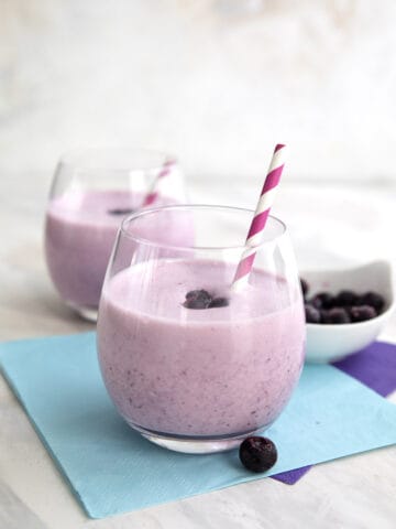 Two keto blueberry smoothies with striped straws sit over purple and blue napkins.