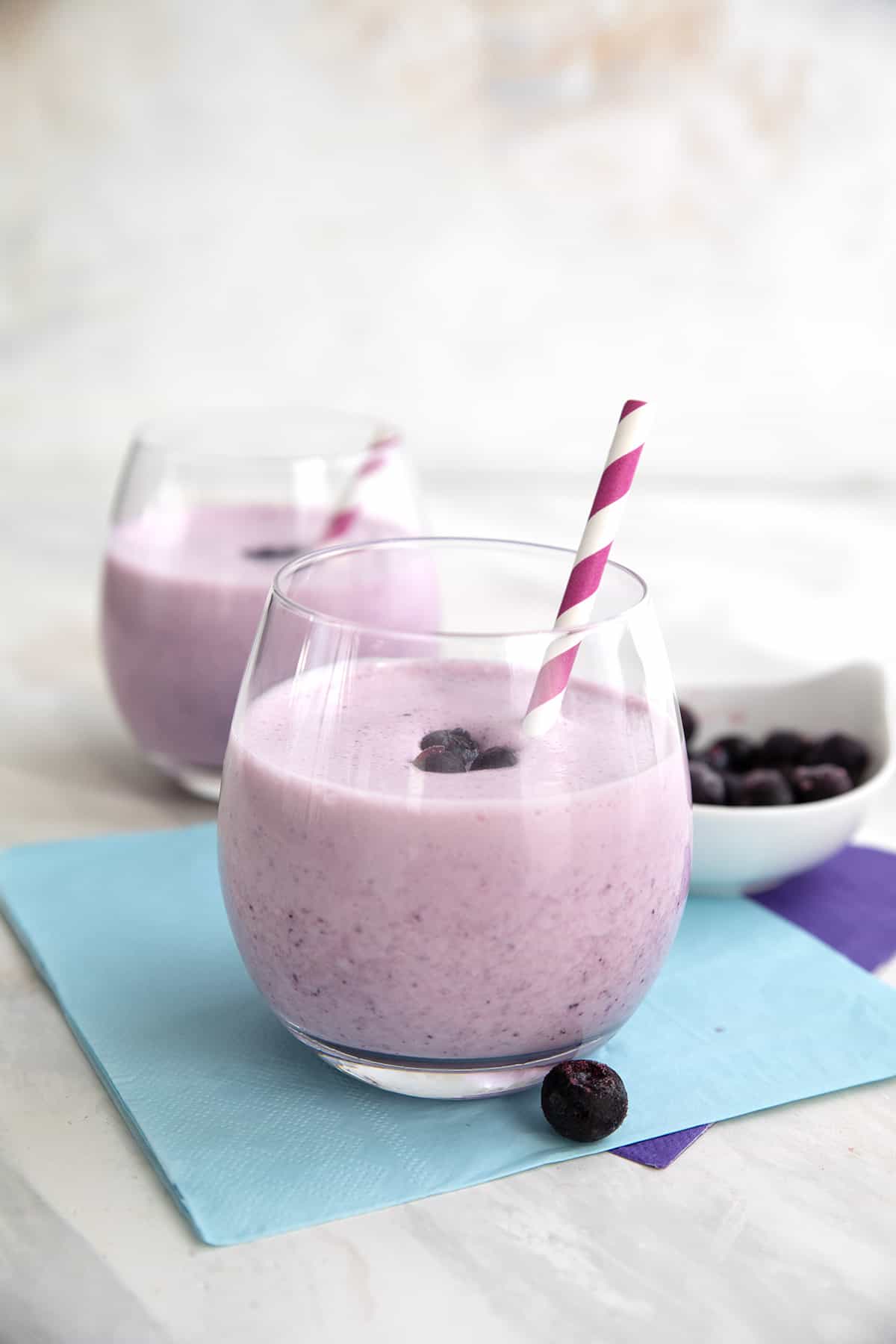 Two keto blueberry smoothies with striped straws sit over purple and blue napkins.