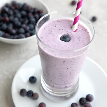 A tall glass filled with keto blueberry smoothie with a bowl of fresh blueberries in the background.