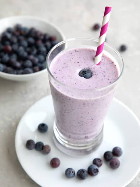 A tall glass filled with keto blueberry smoothie with a bowl of fresh blueberries in the background.