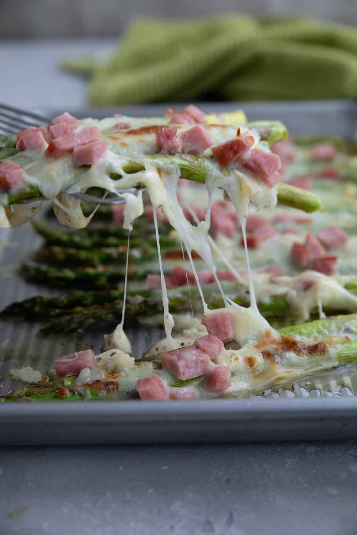 A spatula lifting baked asparagus with melty cheese and ham off of the baking sheet.