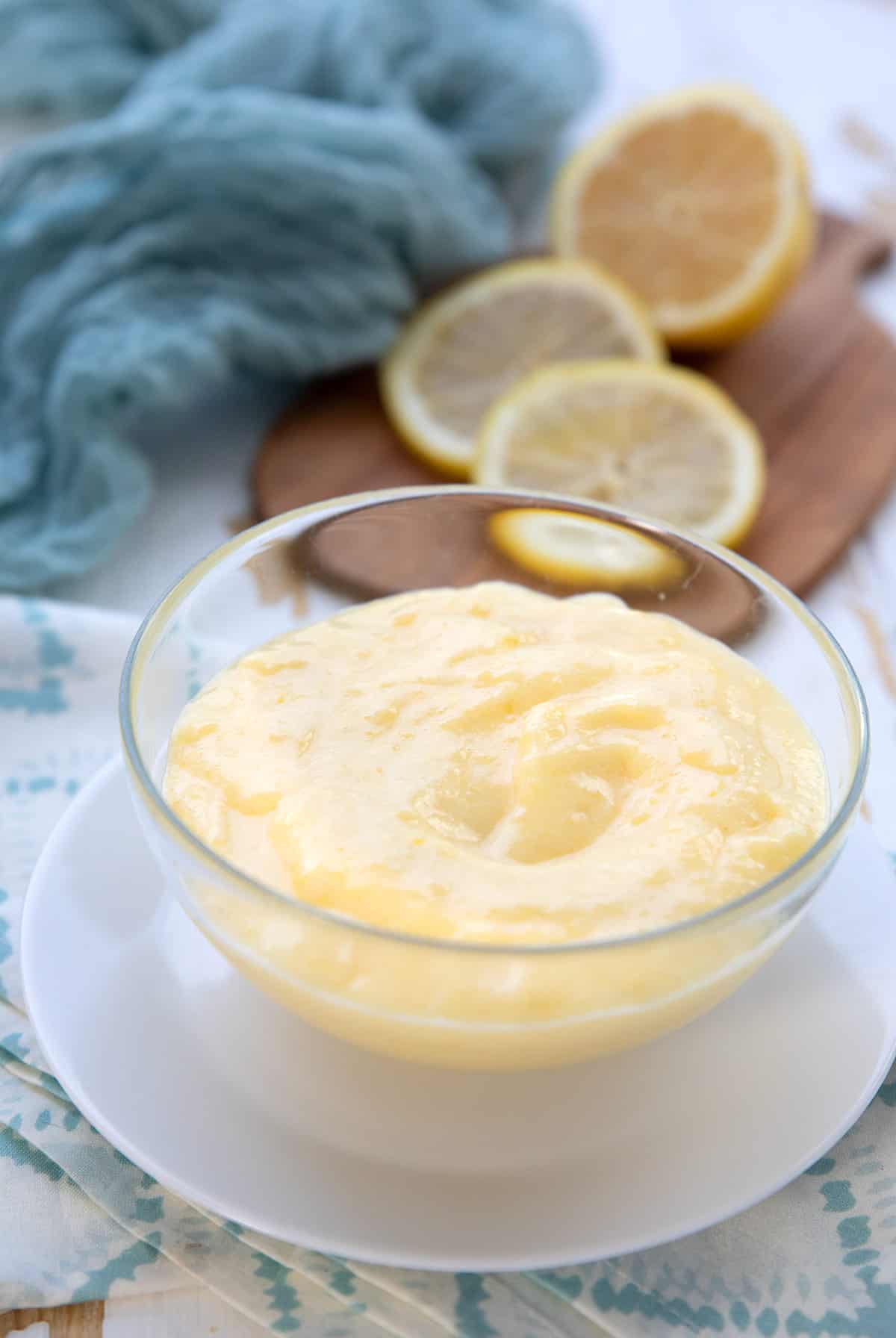A glass bowl filled with sugar free lemon curd with sliced lemons in the background.