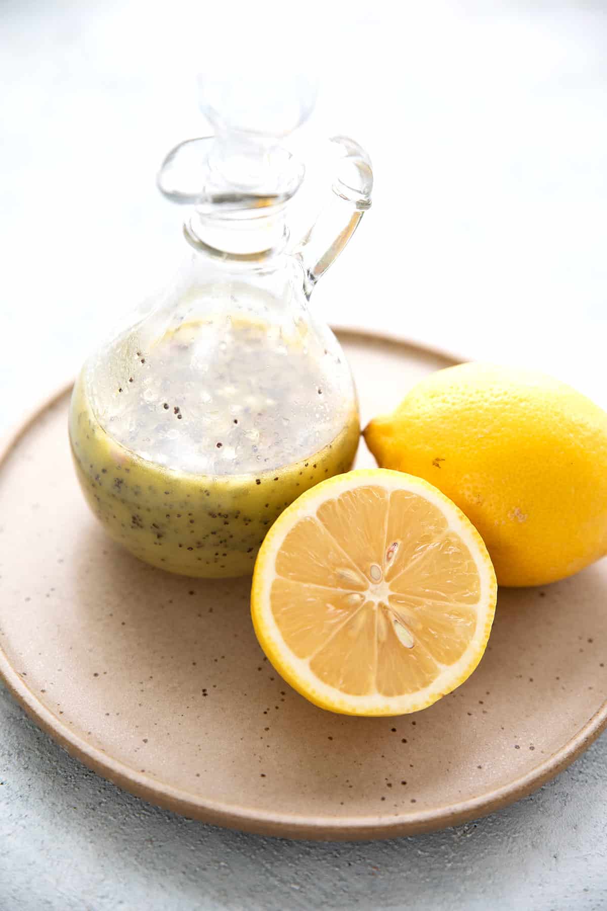 A glass bottle of Keto Lemon Poppy Seed Dressing on a plate with lemons.