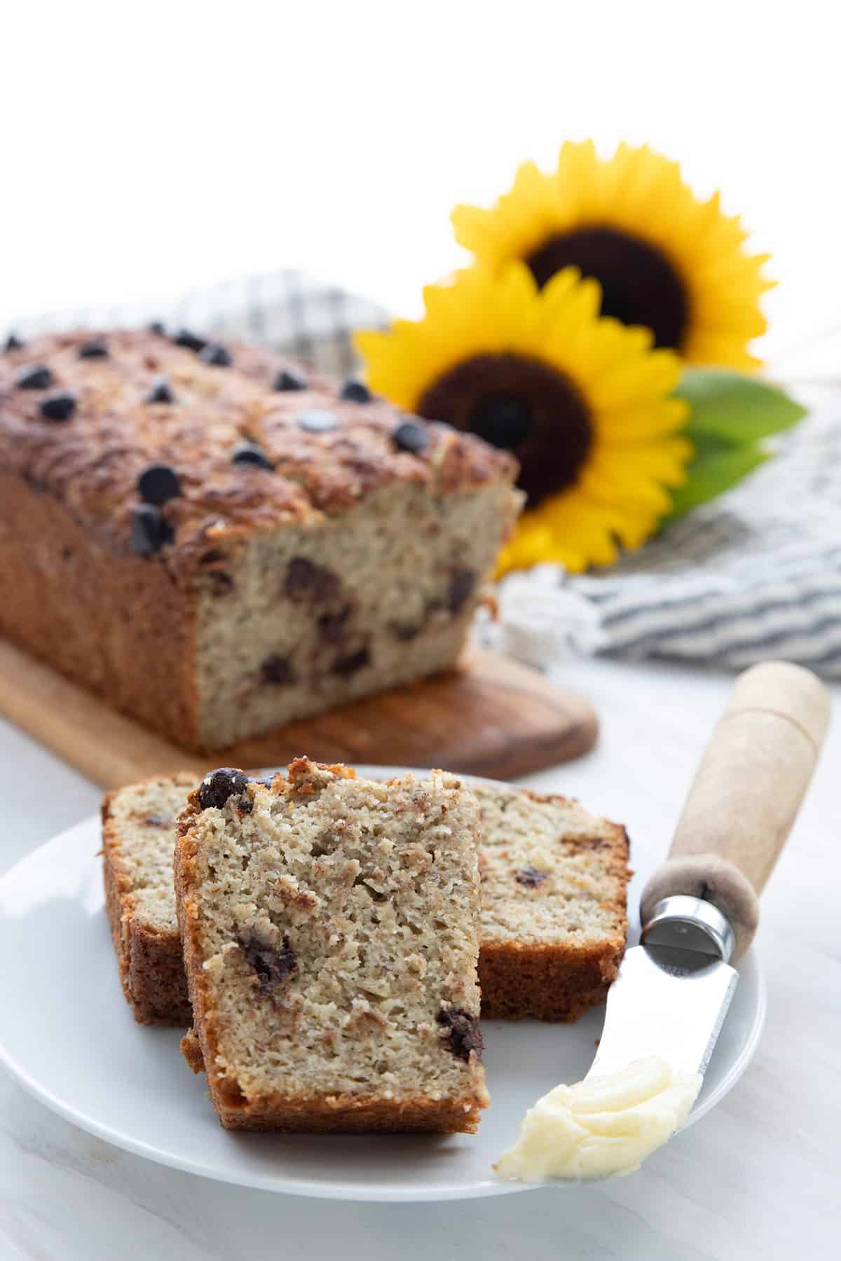 A slice of keto banana bread on a white plate, with the loaf and some sunflowers in the background.