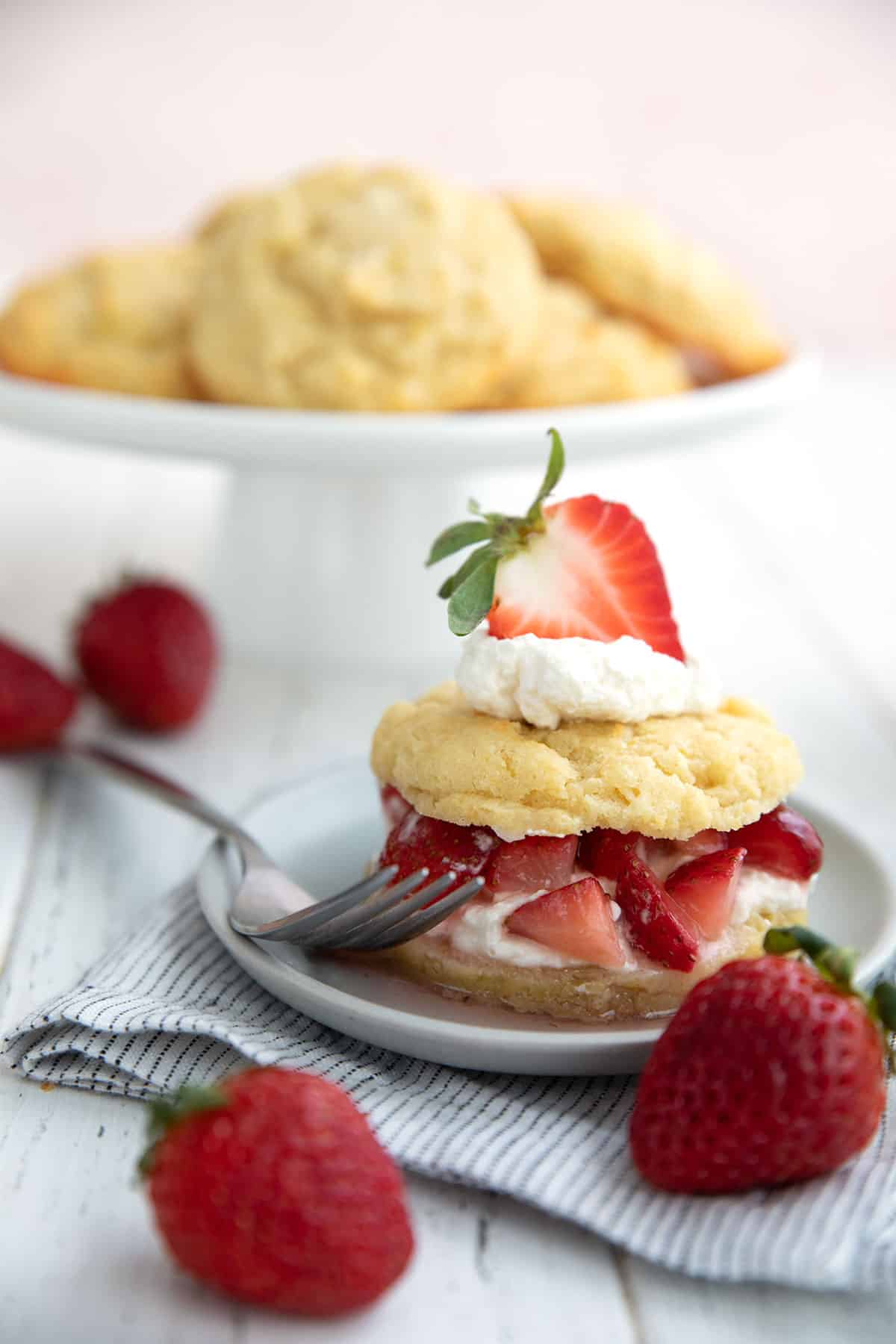 Keto Strawberry Shortcake on a gray plate in front of a stand of biscuits, with strawberries strewn around.