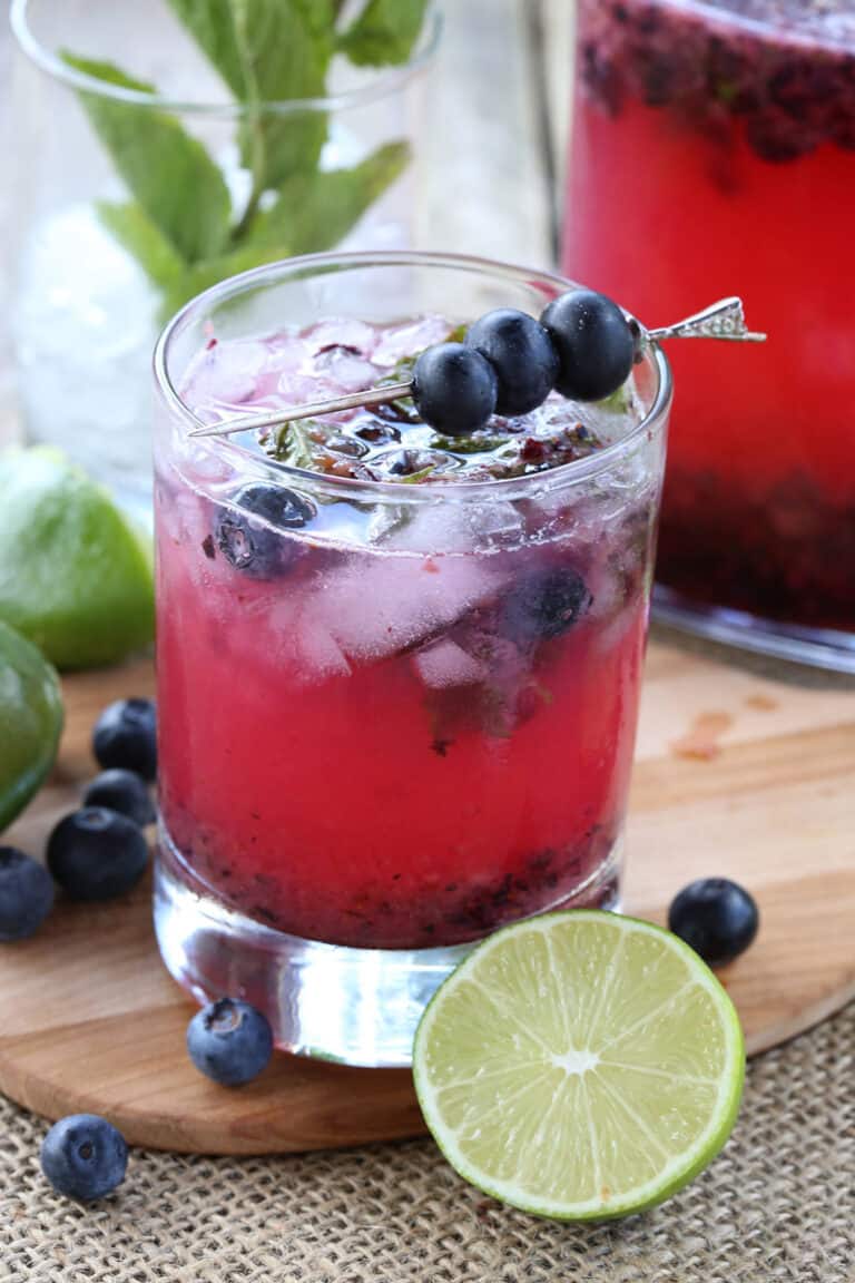 A blueberry keto mojitos on a wooden cutting board with limes, mint, and blueberries around it.