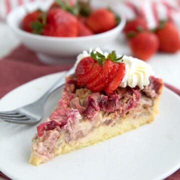 A slice of Rhubarb Custard Pie on a white plate with whipped cream and strawberries on top.