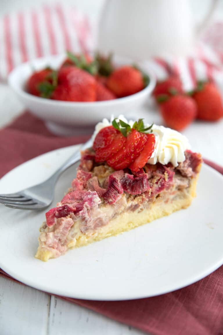 A slice of Rhubarb Custard Pie on a white plate with whipped cream and strawberries on top.